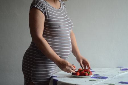 Pregnant woman standing near table with plate of strawberries. Background - white wall. Healthy...