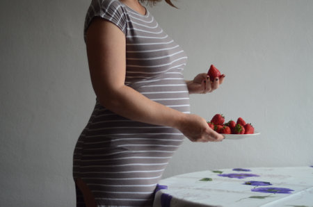 Pregnant woman standing near table with plate of strawberries. Background - white wall. Healthy motherhood food concept, satisfying desires of pregnant womenの写真素材