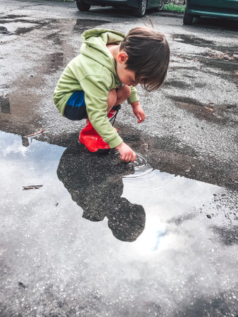 Little boy in a green hoodie and red rubber boots squats near a large puddle after rain, touching the water with his finger. The boy and the sky covered with clouds are reflected in the puddleの写真素材