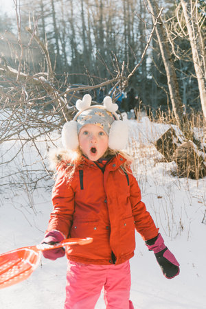 A little girl in a red jacket and earmuffs plays in the snow in the winter forestの写真素材