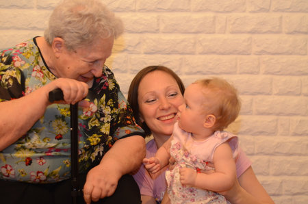 Gray-haired elderly woman with a dog talks to her great-granddaughter. The great-granddaughter is in the arms of her granddaughter. Multigenerational family. Generational relationships. Family day. Background - white brick wallの写真素材