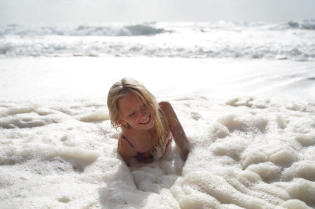 Surreal scene of a shoreline covered in thick, snow-like ocean foam. Little girl, completely smeared with the foam, crawls playfully through the frothy waves. Image captures whimsical and mysterious beauty of nature, blending innocence with the unusual phenomenon of sea foamの写真素材