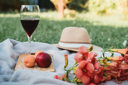 Preparing for a romantic picnic on a green lawn with a glass of red wine, fruits, a straw hat and flowers on a white blanket on a sunny spring dayの写真素材