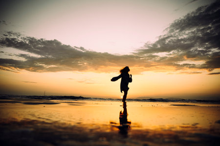 Silhouette of woman joyfully running along the beach at sunset. Golden light reflects on the wet sand, creating a dreamy and peaceful atmosphere. Waves gently touch the shore, and clouds add depth to the sky, enhancing the scene's beauty and serenityの写真素材