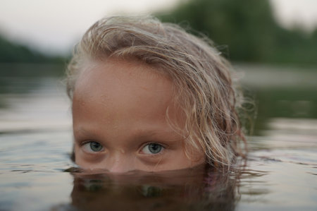 Young girl with wet, wavy hair peers just above the water's surface, her piercing eyes filled with curiosity. Calm natural setting in the background enhances the sense of mystery and tranquility capturing a quiet yet intense moment in natureの写真素材