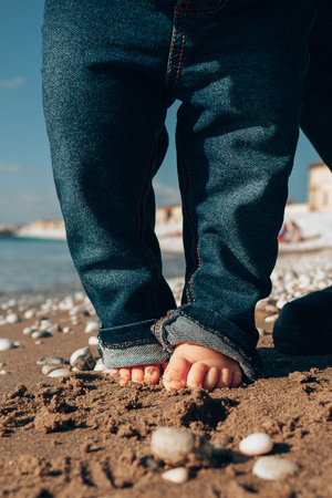 Closeup of a child's barefoot toes peeking out from rolled up jeans as they stand on a sandy beach, capturing a moment of playful interaction with natureの写真素材