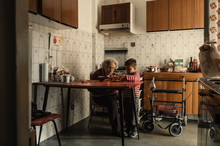 Elderly woman and little child sit together at a table in a homey kitchen, sharing a snack. The atmosphere is filled with warmth and connection, highlighting family bonds. Multigeneration familyの写真素材