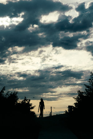 Silhouette of a woman walking on a path during sunset. Dark clouds fill the sky, creating a dramatic and serene atmosphere. Trees frame the sceneの写真素材