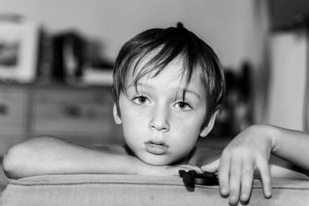 Black and white photo of a child resting their chin on a couch, displaying a thoughtful expression. The setting is indoors with minimal background focusの写真素材