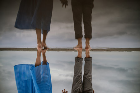 Reflection of two people standing close on the shoreline, showing their feet and legs in casual...