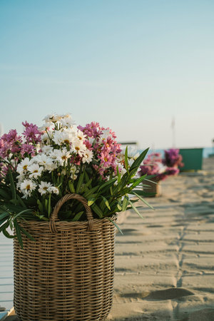 A wicker basket filled with vibrant white and purple flowers placed on a sandy beach. The serene atmosphere suggests relaxation and tranquility by the seasideの写真素材