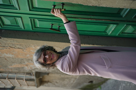 Smiling woman with gray hair wearing glasses and a pink coat opens a green door on an old street....