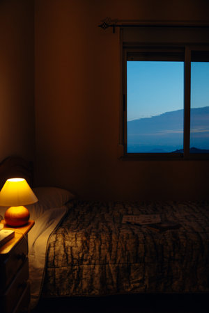 Bedroom scene featuring a neatly made bed next to a warm lamp, with an open window revealing a serene evening sky. Quiet and peaceful ambience, perfect for relaxationの写真素材