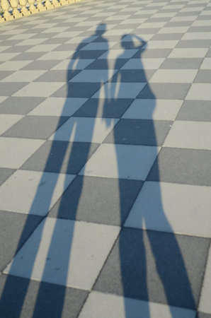 Shadows of two people holding hands are cast on a checkered pavement. The silhouettes create an artistic and abstract depiction of connection and companionship in a public spaceの写真素材