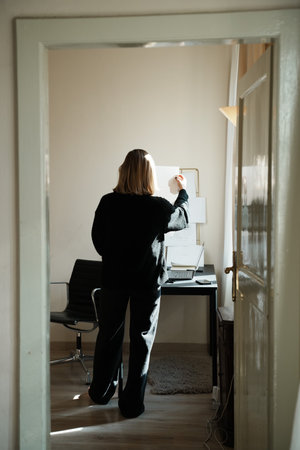 Woman standing at a desk in a home office, analyzing documents. The room is simple with a laptop and chair, indicating a focused work environment. She is looking at paperworkの写真素材