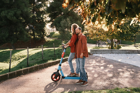 Smiling couple standing together on an electric scooter in a scenic park surrounded by lush...