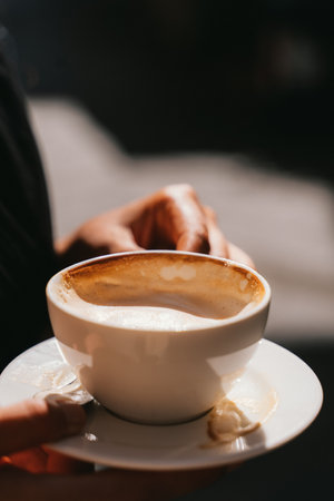 Close-up of hands holding a white cup full of frothy coffee, showing a latte or cappuccino in a simple white dish. The warm light highlights the inviting beverage surface.の写真素材