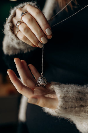 Close-up of a pregnant woman's hands holding a delicate pregnancy harmony ball pendant. Features elegant jewelry with intricately designed silver ring bands and cozy sweater sleevesの写真素材