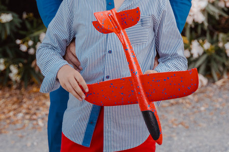 Child dressed in striped shirt and red pants holds a vibrant red toy airplane, embraced by an adult figure. Outdoor scene with a backdrop of green foliageの写真素材