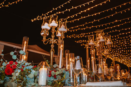 Outdoor dining table adorned with ornate candle holders, glowing candles, and vibrant floral arrangements. Overhead string lights create a festive and romantic atmosphereの写真素材