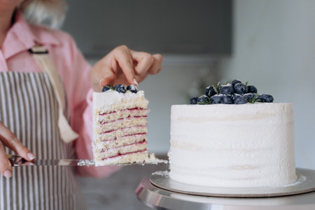 Person on an apron slices a multi-layer cake adorned with blueberries. The cake features alternating layers of vanilla sponge and berry filling. Fresh blueberries top the icingの写真素材