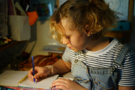 Teen girl focusing intensely on her homework at a desk, holding a blue pen and writing in a notebook. Wearing a striped shirt and denim overalls, creating a studious and dedicated atmosphereの写真素材