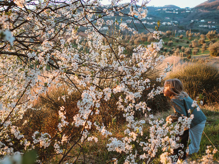 Young girl experiencing the aroma of fresh spring blossoms in a rural landscape. The scene captures the joy of nature and the beauty of blooming trees against a scenic mountain backdropの写真素材