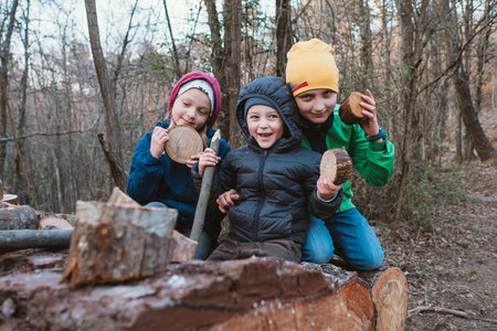 Three children in warm clothing joyfully engage in holding wood slices and sticks in a forest. Their expressions reflect excitement and happiness while exploring nature's wondersの写真素材