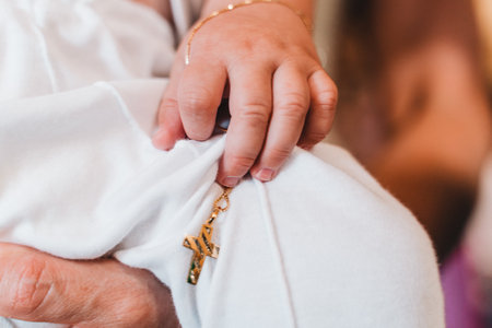 Close-up of a baby's hand gently gripping a gold cross pendant resting on a white fabric. The image...