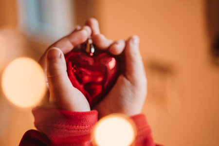 Hands carefully cradle a red heart-shaped ornament, capturing warmth and tenderness. Soft lights surround the scene, creating a cozy atmosphere and highlighting the object's significanceの写真素材
