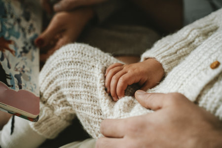 Close-up image of an infant's hand gently resting on an adult's, capturing a tender bonding moment. The soft textures and warmth of the knitted fabric enhance the intimate settingの写真素材