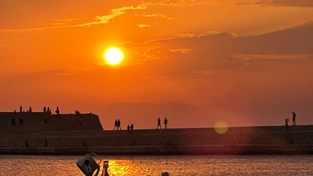 A stunning sunset casts a vibrant orange glow over silhouettes of people standing beside an oceanic stone fort. The serene sea reflects the warm colors, creating a peaceful scene.の写真素材