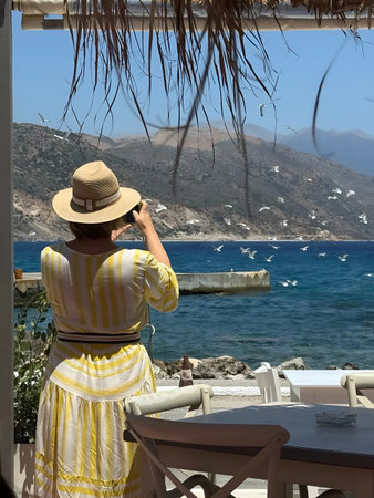 Woman in a yellow dress and straw hat captures a seaside scene on her camera. Seagulls fly over the...