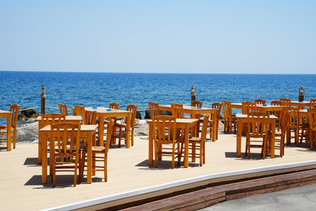 Empty outdoor restaurant terrace with wooden tables and chairs overlooking a calm ocean. The setup is perfect for alfresco dining, offering a serene and inviting atmosphere.の写真素材