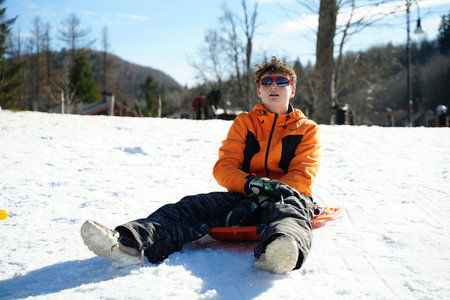 Teen boy wearing an orange jacket sits on a sled, enjoying a sunny winter day. The snow-covered landscape and trees surround him as he experiences the thrill of sledding outdoors.の写真素材