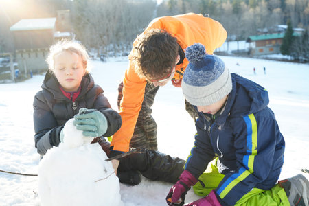 Three children actively engaged in building a snowman on a snowy hillside. Dressed warmly in winter clothes, they work together under bright sunlight, enjoying the winter day.の写真素材