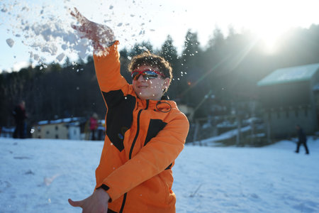 Teen wearing an orange jacket and sunglasses joyfully throws snow into the air during a bright winter day against a backdrop of trees and buildings, showing the fun of snowy activities.の写真素材