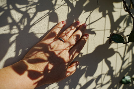 A hand adorned with a ring touches a textured wall, casting intricate shadows of leaves in warm...
