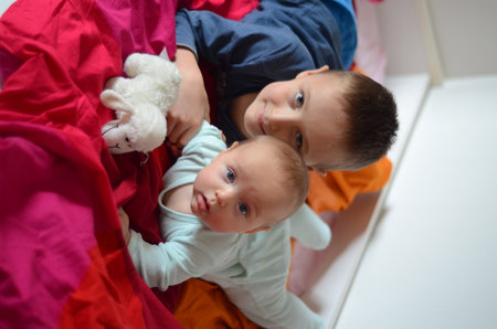 Young siblings lying on a bed enjoying playtime. Older child smiling while the baby curiously looks ahead, both engaged with a toy sheep, highlighting their connection and joyの写真素材