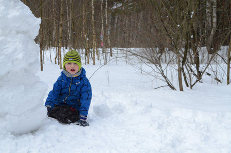 Young child dressed in winter clothes sits happily near a large snowman in a snowy forest. The child...