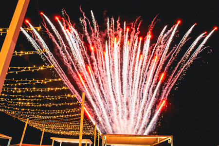 Fireworks burst against the night sky amid festive string lights, creating a vibrant and cheerful atmosphere. The display captures a sense of celebration and joy at a nighttime event.の写真素材