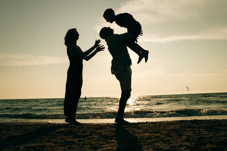 A joyful family moment captured at the beach during sunset. A parent playfully lifts a child while another reaches out, creating a silhouette against the glistening sea and sky.の写真素材