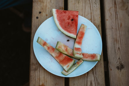 Watermelon rinds left on a plate after being eaten outdoors. The remnants reveal the enjoyment of a refreshing snack on a wooden table, highlighting summertime relaxation.の写真素材