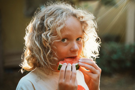 Young child with curly hair enjoying a slice of watermelon outdoors. The image captures a moment of simple pleasure and summer freshness, highlighting a joyful and playful mood.の写真素材