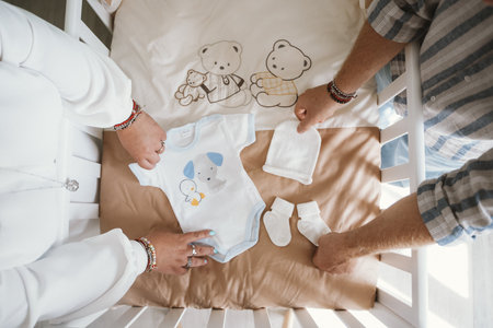 Parents arranging baby clothing and accessories in a crib, including a onesie, hat, and socks. The scene captures anticipation and care in preparing for a baby's arrival.の写真素材