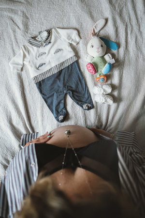 Pregnant woman rests hand on belly while looking at baby clothes and toys on bed, creating a warm and hopeful scene focused on the upcoming arrival of a newborn.の写真素材