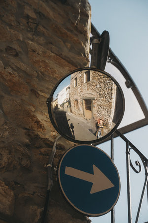 Street mirror reflects a quaint town street with rustic stone buildings and a blue traffic sign pointing right. A person appears in the reflection, contributing to the street's charm.の写真素材