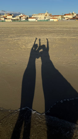 Two long shadows of people standing on a sandy beach holding hands. The sunlight creates distinct silhouettes against the sand, capturing a moment of connection and playful gesture.の写真素材