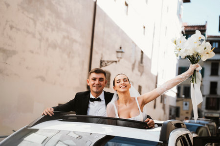 Newlywed couple celebrating their wedding day by standing through the sunroof of a car. The bride joyfully raises her bouquet with a bright smile, while the groom looks happily at the camera.の写真素材