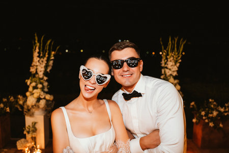 A couple celebrates their wedding joyfully, wearing playful heart-shaped sunglasses that display their special date. Their expressions radiate happiness and excitement in a festive atmosphereの写真素材